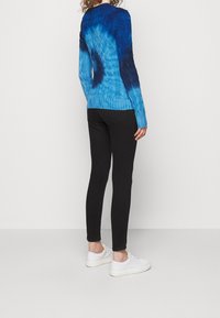 Blå och marinblå tie-dye stickad tröja med ribbad fåll, matchad med svarta tajta byxor och vita sneakers. Sedd bakifrån.