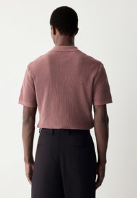 Korte mouwen, geribd poloshirt in de kleur dusty rose. Heeft een zachte textuur en een kraag, gecombineerd met op maat gemaakte zwarte broek.