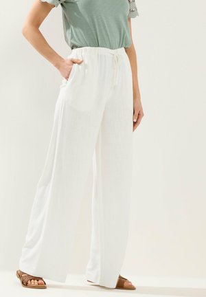 Kobieta ubrana w luźne białe spodnie z troczkiem, zielony krótki rękaw i brązowe plecione sandały, stojąca z jedną ręką w kieszeni.