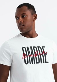 Camiseta blanca de algodón con la impresión negra prominente "OMBRE" y acentos en cursiva rojos en el centro. Diseño clásico de cuello redondo.