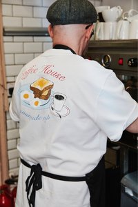 Mężczyzna w białej koszuli z grafiką śniadaniową i napisem, pracujący przy ekspresie do kawy w kuchni z białymi, kafelkowymi ścianami.