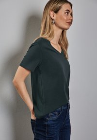 Tmavě zelené tričko s výstřihem do V a krátkými rukávy, vyrobené z hladké látky. Nosené s tmavě modrými džínami, které mají mírně zvýrazněný boční lem.