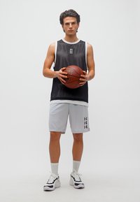 Νεαρός άνδρας με μαύρο αμάνικο μπλουζάκι και λευκό σορτς κρατάει μια μπάλα μπάσκετ, στέκεται μπροστά σε ένα απλό φόντο.