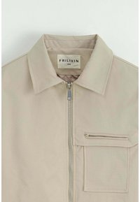 Veste zippée beige avec un col, une poche avant et une poche poitrine zippée. Fabriquée en tissu lisse, elle présente des coutures visibles et une étiquette de marque.