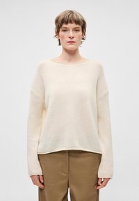 Jasnobeżowy sweter o luźnym kroju, z szerokim dekoltem i długimi rękawami. Tkanina wydaje się miękka, z delikatną fakturą i nieco dłuższym tylnym brzegiem.