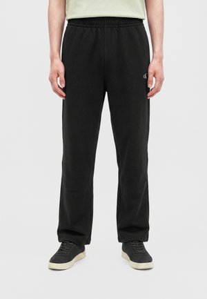 Personne vêtue d'un pantalon de jogging noir Calvin Klein avec une taille élastique et des baskets noires aux semelles blanches, debout devant un fond gris.