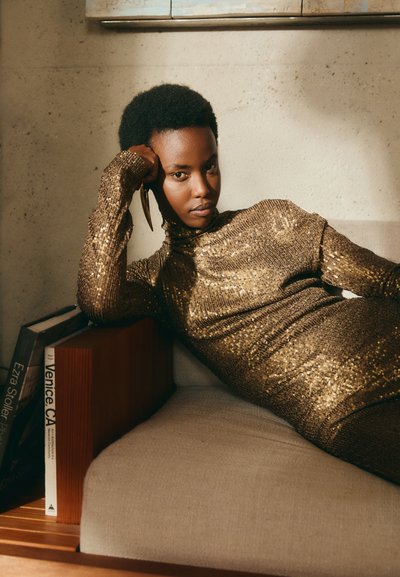 Robe en sequins dorés avec un col haut et des manches longues. Le mannequin est allongé sur un canapé de couleur claire à côté de livres. Texture lisse.