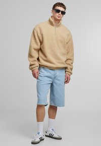 Pull en polaire beige avec col zippé mi-haut, associé à un short en jean bleu clair. Le mannequin porte des chaussettes blanches et des baskets noir et blanc.