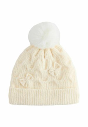 Bonnet en tricot crème avec un pompon blanc duveteux sur le dessus, présentant un motif en câble et trois petits nœuds sur le côté.