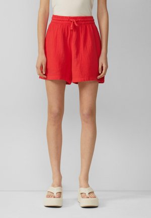 Person iført lyse røde shorts med snøre og beige platformsandaler, der står mod en neutral baggrund.