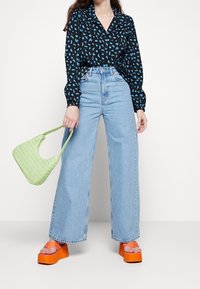 Blusa nera con stampa leopardata blu, jeans a vita alta azzurri a gamba larga, sandali con plateau arancioni e borsa intrecciata verde.