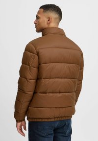 Brun puffjakke med kviltet tekstur, høy krage, elastiske mansjetter og hem. Båret med mørkeblå jeans, vist fra baksiden.