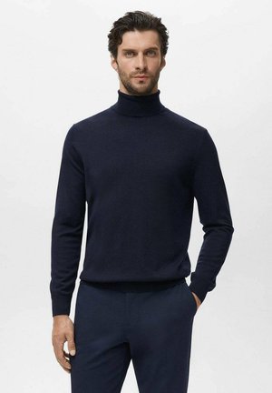 Hombre con cabello oscuro rizado, vistiendo un suéter de cuello alto azul marino y pantalones a juego, de pie con una mano en el bolsillo.