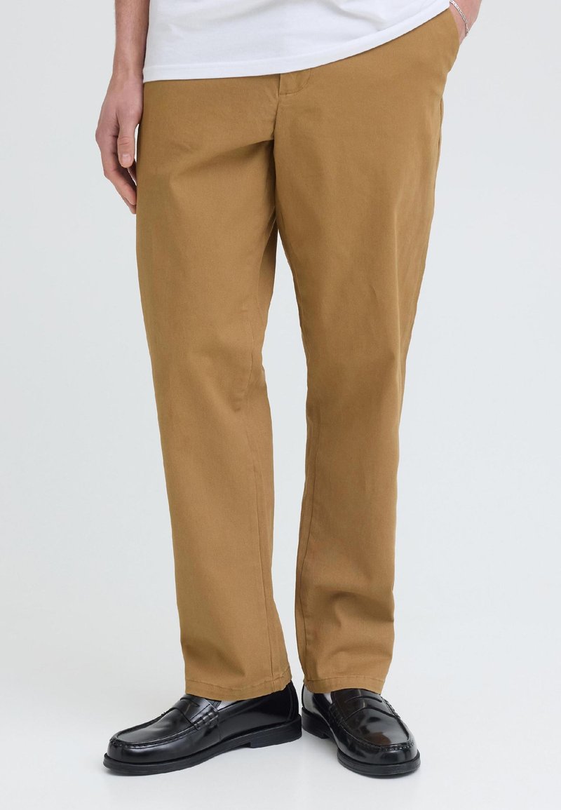 Homme portant un pantalon marron, des mocassins en cuir noir et une chemise blanche, debout avec une main dans la poche sur un fond clair uni.