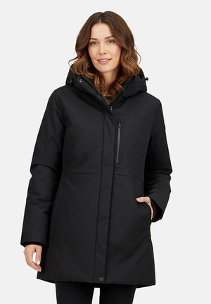 Vrouw draagt een zwart midilengte winterjas met capuchon, ritssluiting op de borstzak en hand in de zijzak, lachend naar de camera.