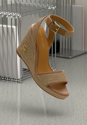 Tanfarvet flettet kilehæl sandal med ankelstrop, guldbeslag og Tommy Hilfiger-logo på kilehælen, vist mod en minimalistisk baggrund.