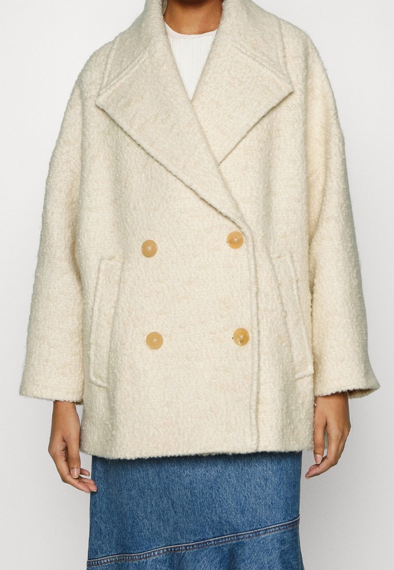 Manteau à double boutonnage en laine texturée écru avec de larges revers, boutons marron clair et poches latérales, porté par-dessus une jupe en denim.