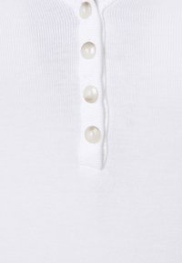 Débardeur blanc côtelé avec une courte patte de boutonnage dotée de quatre boutons transparents. Le tissu semble doux et léger.