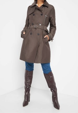 Kobieta w brązowym, dwurzędowym płaszczu z paskiem, niebieskich dżinsach i ciemnobrązowych, teksturowanych kozakach za kolano, stojąca z rękami w kieszeniach.