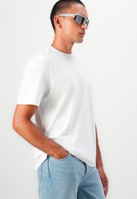 Weißes T-Shirt aus Baumwolle mit kurzen Ärmeln und Rundhalsausschnitt, versehen mit einem dezenten Logo. Kombiniert mit hellblauen Jeans und grauen Sonnenbrillen.