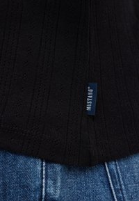 Nahaufnahme eines schwarz strukturierten Stoffes mit einem kleinen blauen Mustang-Label über blauen Jeans.