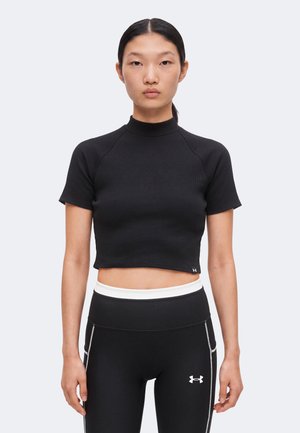 Νεαρή γυναίκα με μαύρο crop top με κοντά μανίκια και μαύρο αθλητικό κολάν με ψηλή μέση, στέκεται μπροστά σε απλή φόντο.