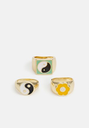 Três anéis de ouro com designs yin-yang; um com esmalte preto e branco, um em formato quadrado com detalhes em verde menta e um com um padrão de flores amarelas.