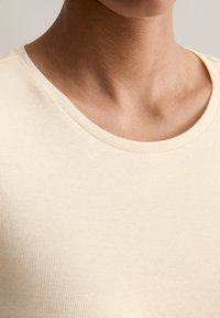 Hellbeige geripptes Baumwollshirt mit Rundhalsausschnitt, das eine glatte Textur und subtile Nähte zeigt. Die Nahaufnahme hebt die Stoffdetails hervor.