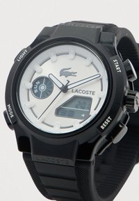 Μαύρο ρολόι Lacoste με άσπρη πρόσοψη, αναλογικές δείκτες, ψηφιακή οθόνη, ένδειξη ημέρας και ετικετοποιημένα κουμπιά: φως, έναρξη, λειτουργία, επαναφορά.