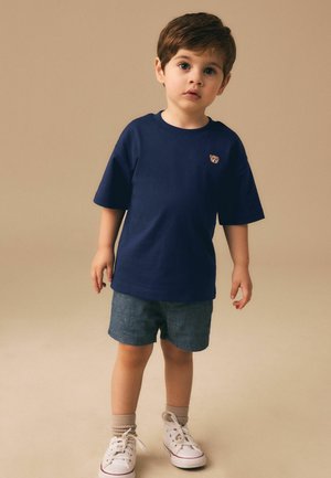 Ung dreng iført en marineblå t-shirt med et lille bamsepatch, denimbukser, beige sokker og hvide sneakers, stående på en neutral baggrund.