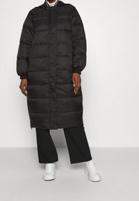 Sort pufferjakke med vandret syning, hætte og lange ærmer. Parret med sorte bukser med brede ben og hvide sneakers på en neutral baggrund.