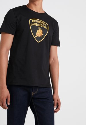 Man draagt een zwart T-shirt met goudkleurig Automobili Lamborghini-logo en donkerblauwe spijkerbroek tegen een effen lichte achtergrond.