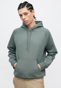 Duks s kapuljačom u prigušenoj zelenoj boji, napravljen od mekanog pamuka, s prednjim džepom, rebrastim manšetama i malim logotipom na rukavu.