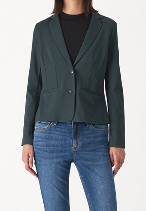 Femme portant un blazer vert foncé cintré sur un haut noir, associé à un jean denim bleu, debout devant un fond uni.