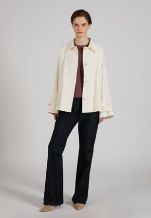 Young woman standing wearing a cream buttoned jacket, maroon top, dark wide-leg pants, and beige pointed shoes against a plain gray background.