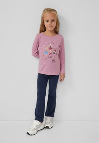 Langärmliges, pinkes Hemd mit verspieltem Grafikdesign, das bunte Figuren zeigt; kombiniert mit dunkelblauen Hosen und weißen Sneakers.