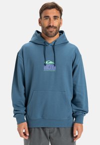 Un hoodie bleu en matériau doux avec une poche avant. Le design comprend un graphique du logo Quiksilver en vert clair et violet.