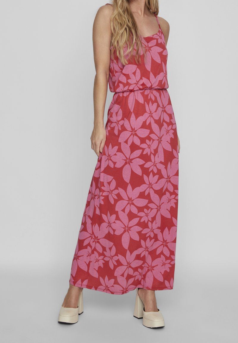 Maxi dress in red fabric featuring a pink floral pattern, with a sleeveless design and an elastic waist. Model wears white platform shoes.