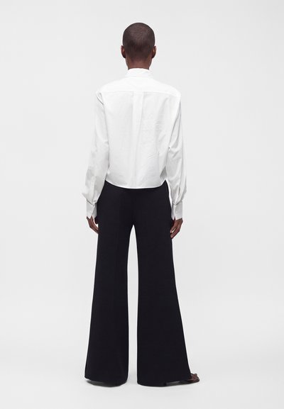 Personne debout de dos portant une chemise blanche à manches longues et un pantalon noir à jambes larges sur un fond clair uni.