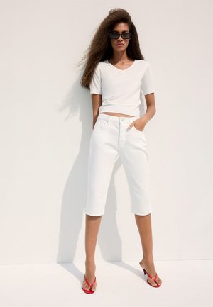 Femme aux longs cheveux bouclés portant des lunettes de soleil noires, un haut blanc à manches courtes, un pantalon blanc jusqu'aux genoux et des sandales rouges à talons, debout contre un mur.