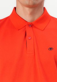 Rød polo t-shirt lavet af åndbart stof med klassisk krave, to-knaps placket og et lille, mørkt logo på venstre side.