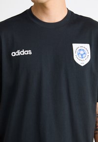 Fekete pamut póló, kerek nyakkivágással, hímzett fehér "adidas" logóval, valamint fehér folttal, kék focis grafikával és szöveges részletekkel.