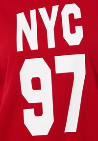 Camiseta roja con letras en bloque blancas y audaces que dicen "NYC 97". La tela parece suave con un diseño simple.