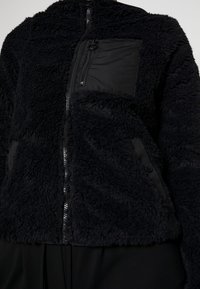 Giacca in pile nera con un esterno testurizzato e soffice, chiusura anteriore con zip e una tasca liscia a contrasto con dettaglio zip.