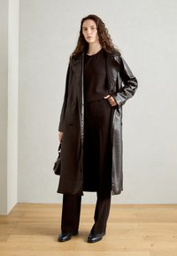 Larga gabardina de cuero negra sobre una blusa negra y pantalones de pierna ancha, combinada con zapatos negros y un bolso pequeño. Textura suave, corte ajustado.