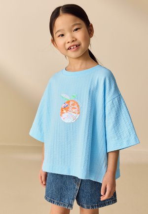 Ung pige smilende, iført en lyseblå tekstureret skjorte med et orange frugtmotiv og denimshorts, stående mod en beige baggrund.
