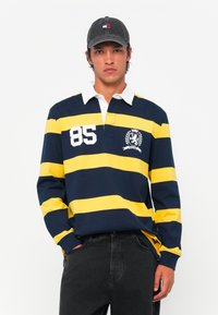 Jūrinės tamsiai mėlynos ir geltonos juostelės ilgomis rankovėmis polo marškiniai su balta apykakle, puošti liūto emblema ir skaičiumi "85". Sukomplektuota su tamsiai mėlynomis džinsomis.
