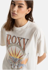 Biała bawełniana koszulka z okrągłym wzorem przedstawiającym słońce, deskę surfingową i skomplikowane wzory. Wyraziste napisy "ROXY" w kolorze pomarańczowym.