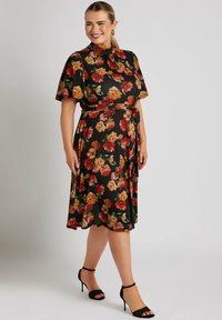 Vestido floral con fondo negro, mangas cortas, cintura ceñida y falda a la altura de la rodilla, con rosas naranjas y rojas, combinado con tacones negros.
