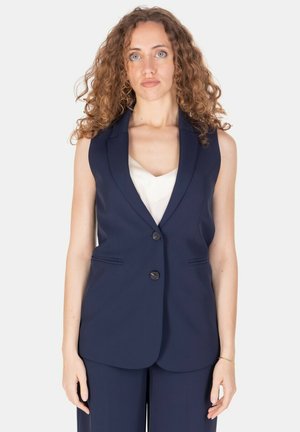 Donna con capelli ricci che indossa un blazer blu navy senza maniche e pantaloni abbinati, in piedi davanti a uno sfondo bianco semplice.